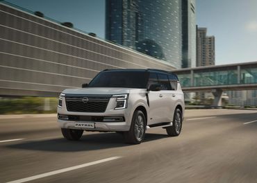 A white Nissan Patrol SUV driving on a city road with modern buildings in the background.