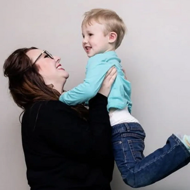 A joyful woman lifts a smiling toddler against a plain background.