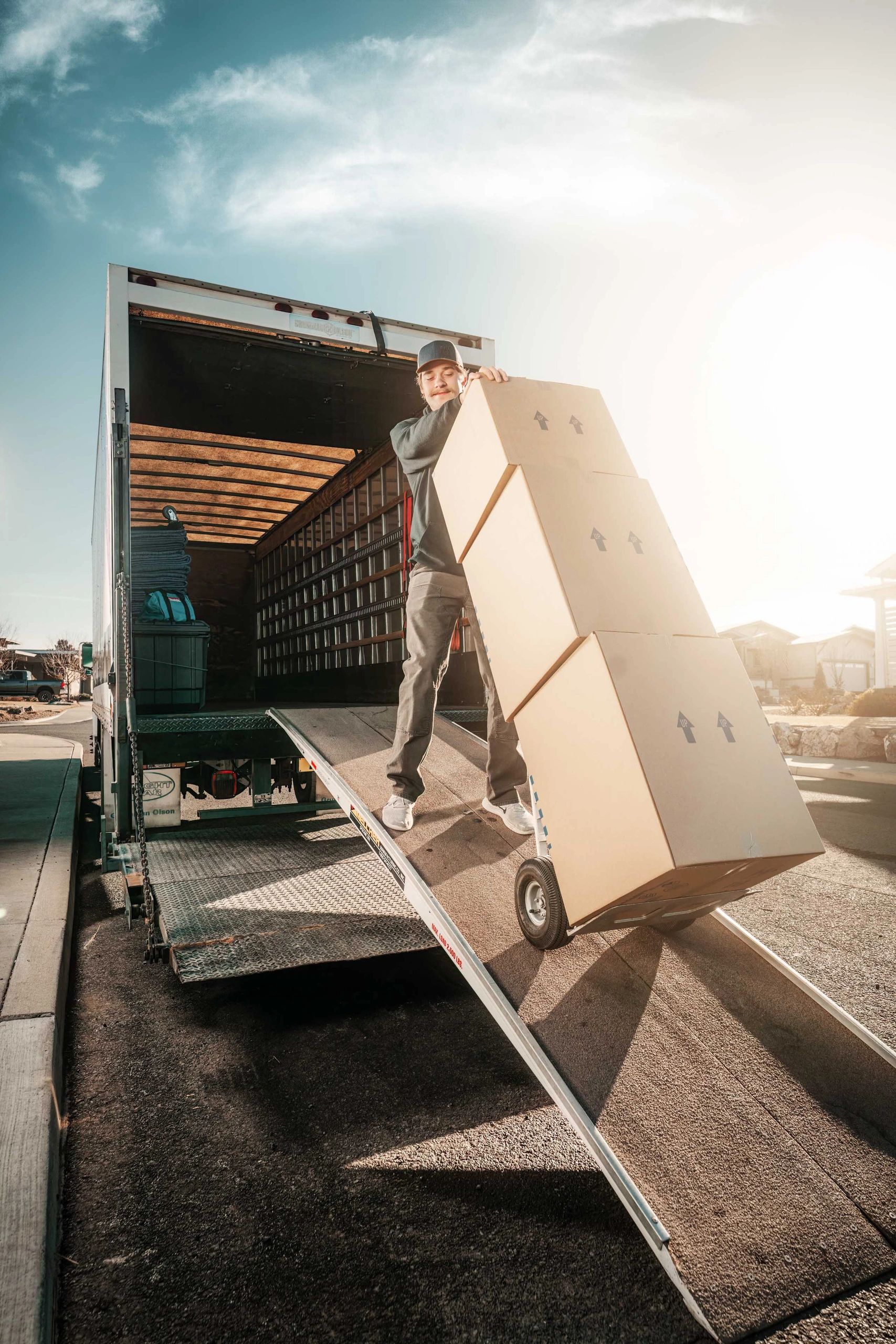 A mover is walking down the ramps of a moving truck while pushing a handtruck brimming with boxes.