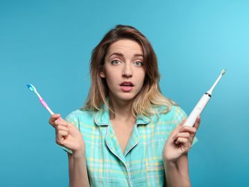 Woman holds electric toothbrush in left hand and manual toothbrush in right hand with puzzled look