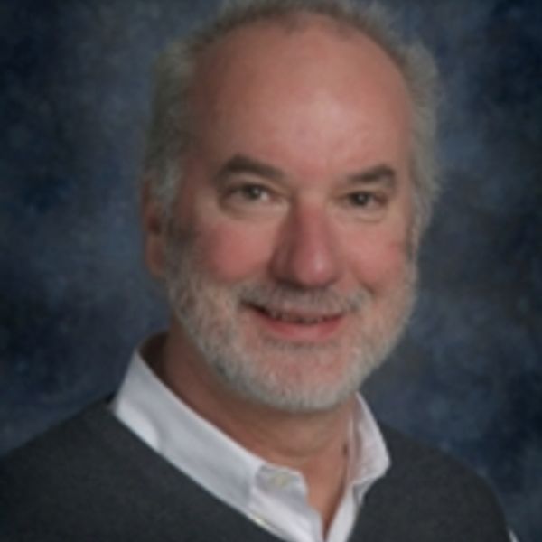 A smiling older man with a white beard and light hair against a blue background.