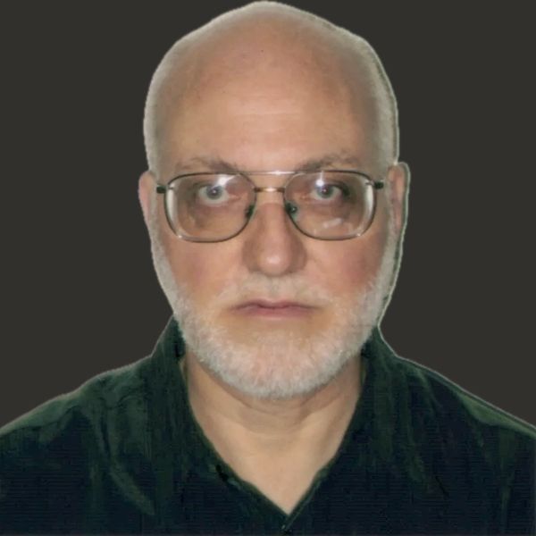 Portrait of a serious elderly man with glasses and a white beard against a dark background.