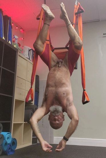 Man practices aerial yoga inverted in an orange hammock in a studio.
