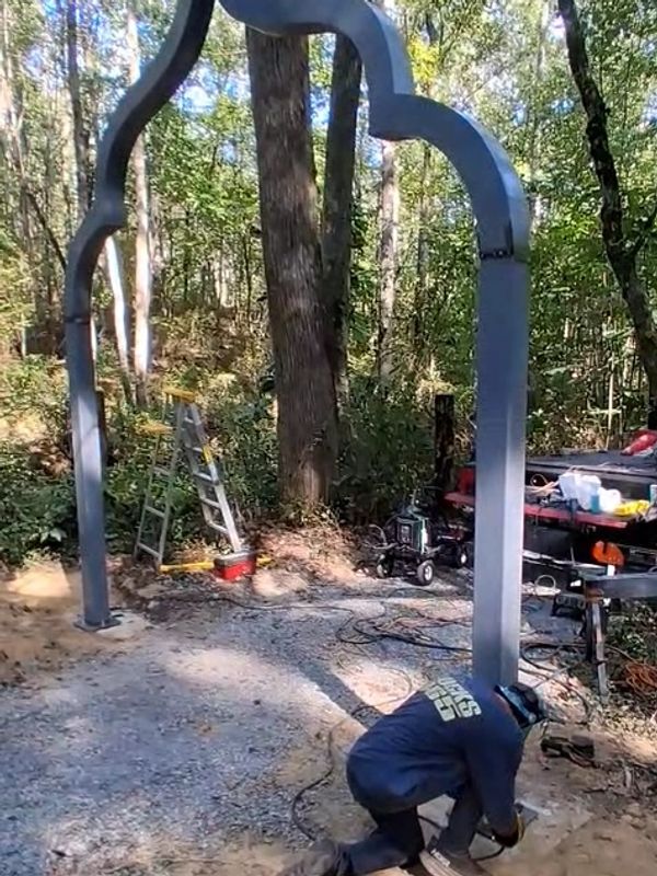 Person welding metal arch in forested area.