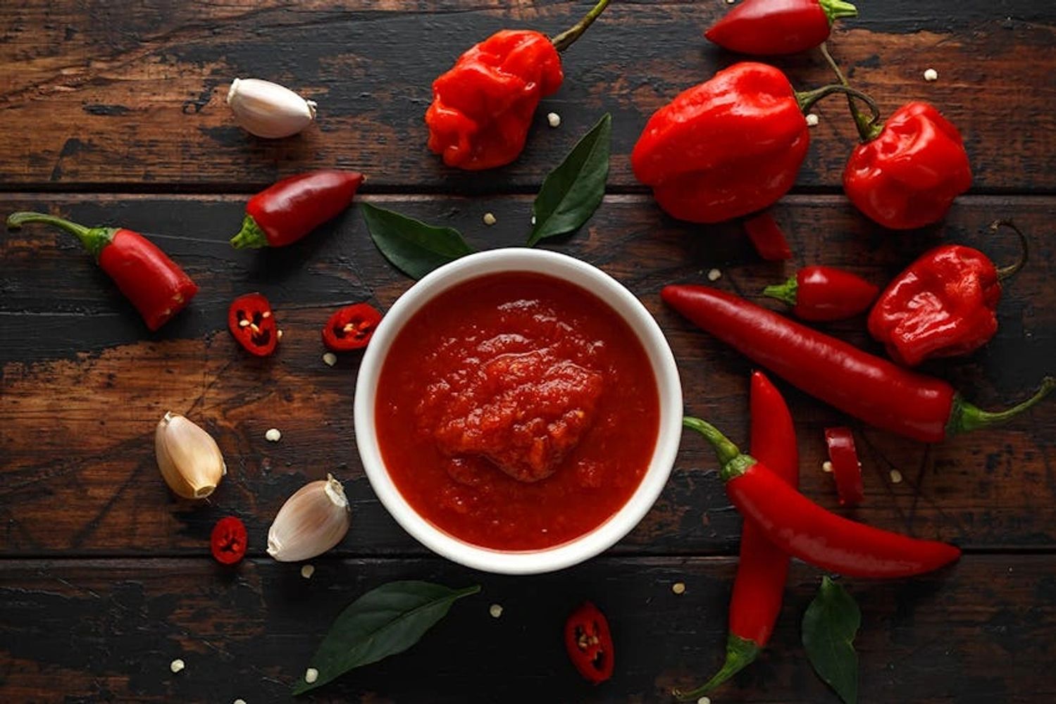 A bowl of red chili sauce surrounded by various red chili peppers and garlic cloves.