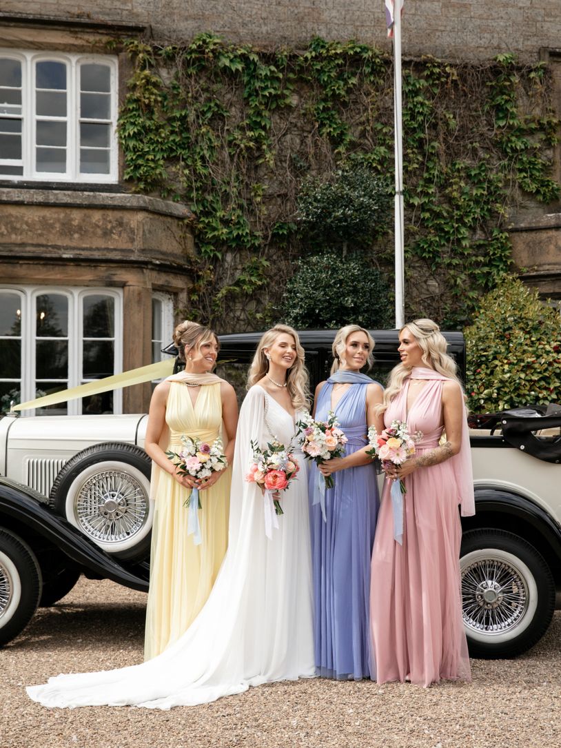 Four women in pastel dresses holding bouquets pose by a vintage car.