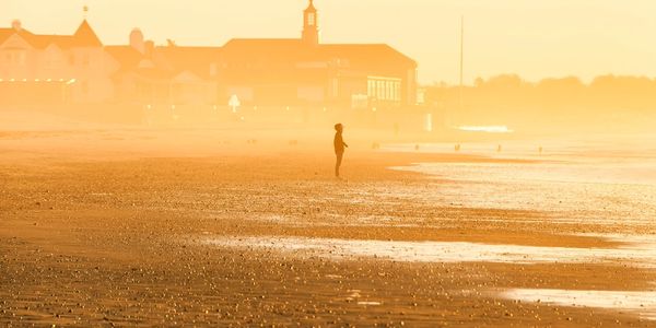 A person stands alone on a misty beach with buildings in the background at sunrise or sunset.