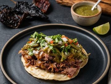 A delicious taco topped with shredded meat, guacamole, onions, and cilantro on a black plate.