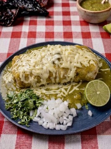 Chimichanga covered in green sauce and melted cheese with lime, onions, and cilantro on the side.