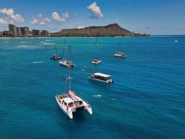 Sailboats and snorkelers in clear blue ocean near a city and mountain.