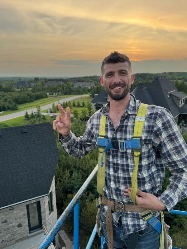 Man in safety harness smiles and gestures atop a building at sunset.