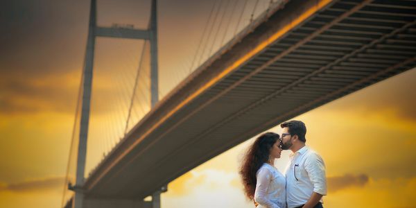 silhouetted couple standing near a river, with a suspension bridge and a golden sunset in the back