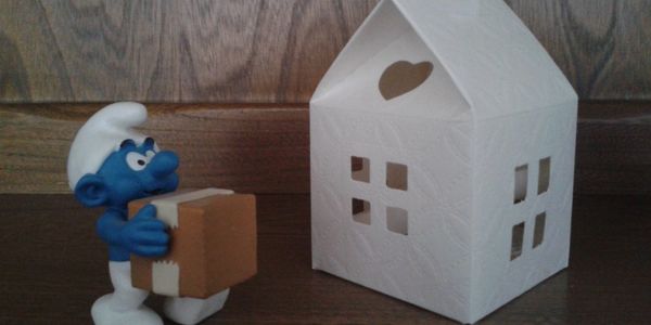 Un Schtroumpf tenant une boîte devant une maison blanche en papier.