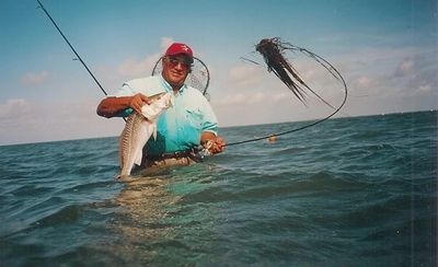 Man standing in water holding a fish and fishing rod with seaweed tangled on the line.