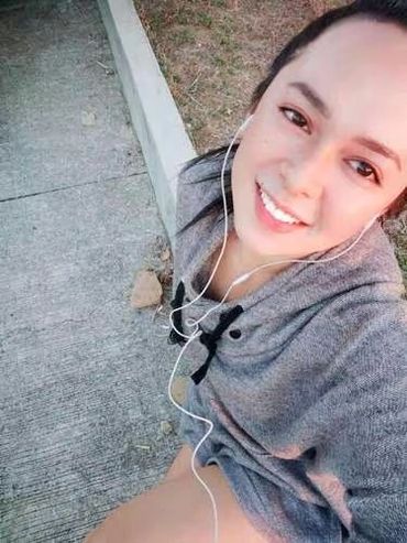 A smiling woman wearing earphones and a gray hoodie sitting on a sidewalk.