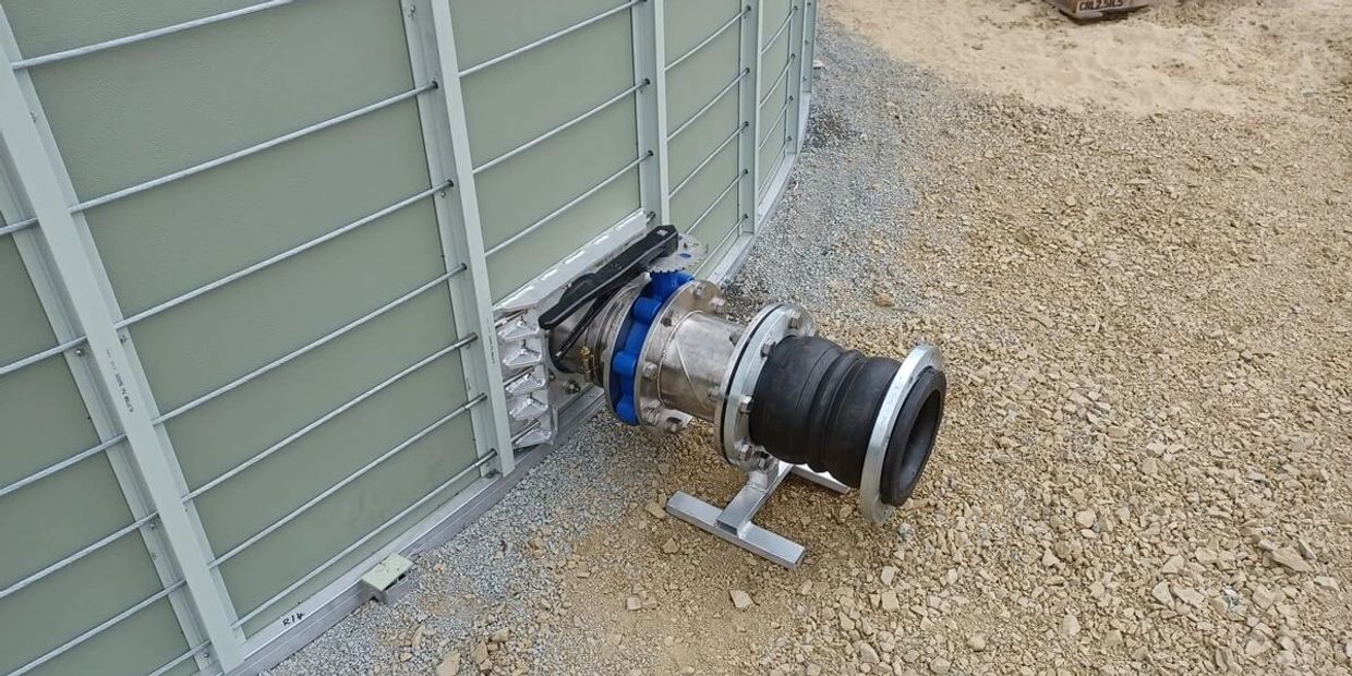 Industrial pipe connection with valve on gravel ground next to a metal wall.