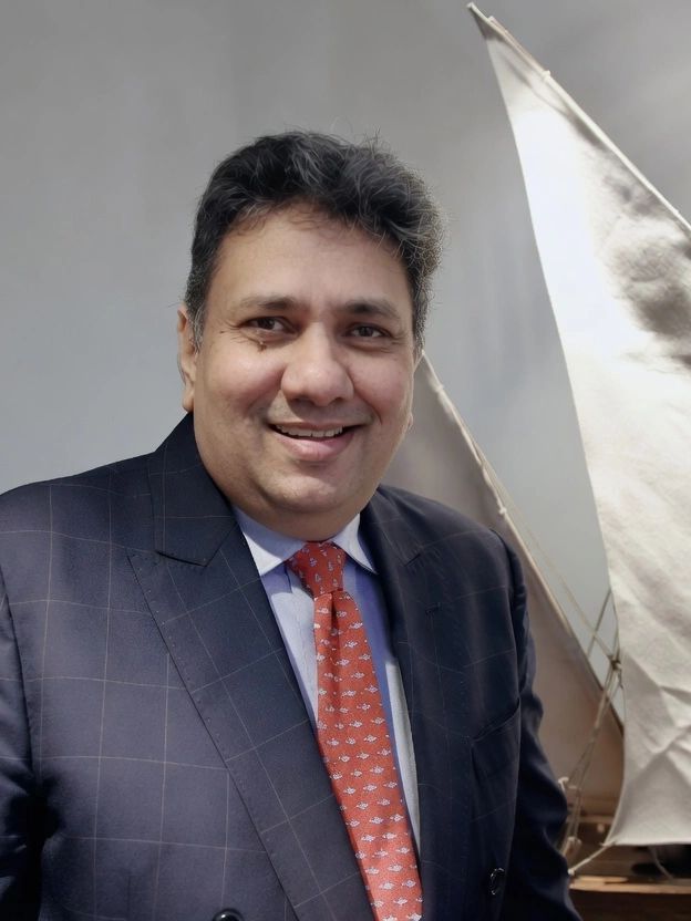 A smiling man in a suit and red patterned tie stands by a model sailboat.