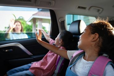 Two children in a car waving goodbye to a woman outside.