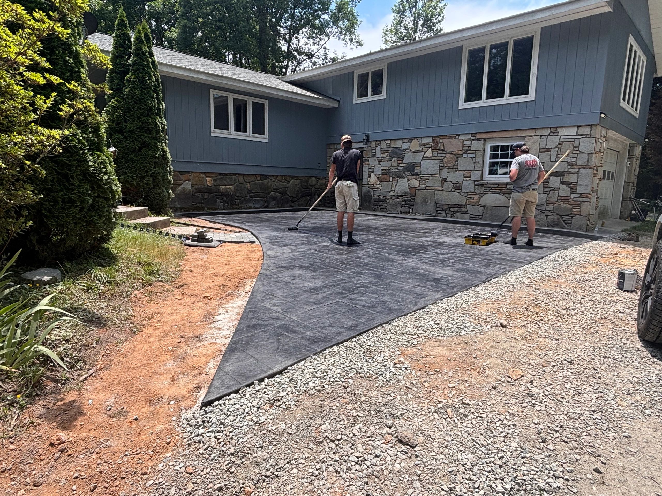 Two men working on a newly paved driveway outside a blue house.