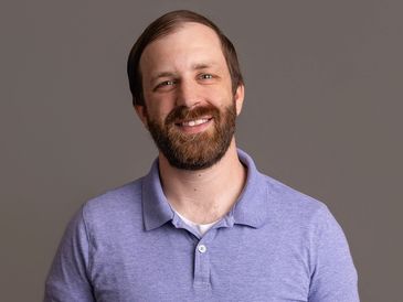 Smiling man with a beard in a purple polo shirt.