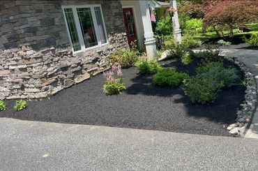 Front yard garden with black mulch and green shrubs by a stone house.