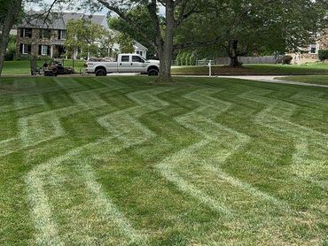 Freshly mowed lawn with zigzag patterns under large trees and a parked truck.