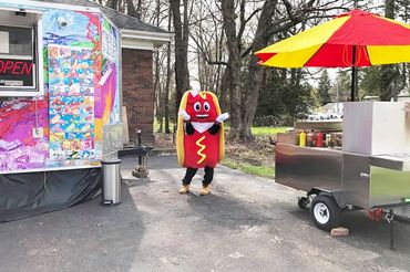 Person in hot dog costume dancing between food stands outdoors.
