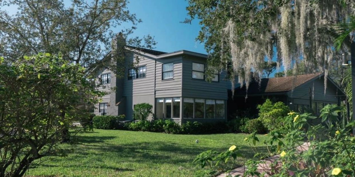 A two-story house surrounded by lush greenery and trees under a clear blue sky.