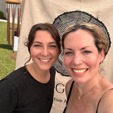 Two women smiling in front of a decorative backdrop outdoors.