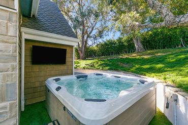 Outdoor hot tub next to a house with a mounted TV and greenery.