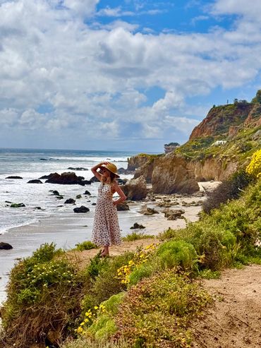 A woman in a floral dress stands on a cliff overlooking the ocean.