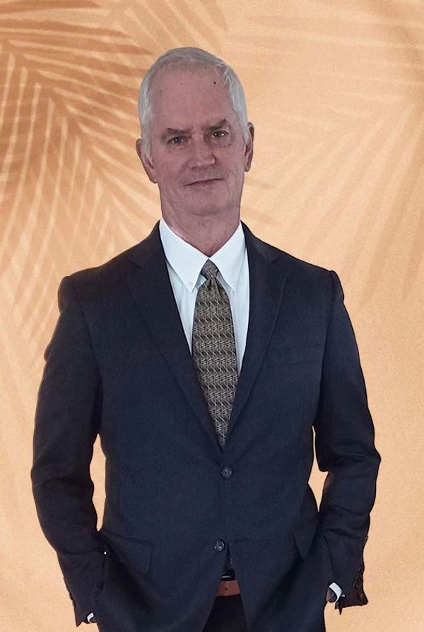 Man in a dark suit and patterned tie posing against a beige backdrop with palm leaf shadows.