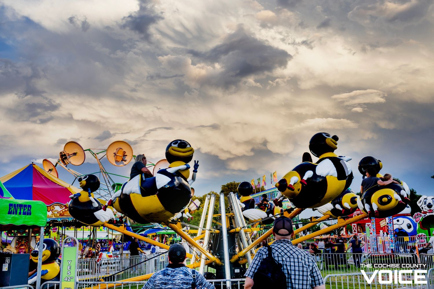 Families enjoy the bee-themed carnival ride under a dramatic cloudy sky.