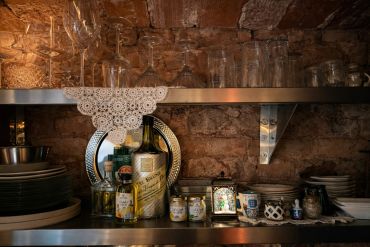 Rustic kitchen shelf with bottles, plates, and glassware against a brick wall.