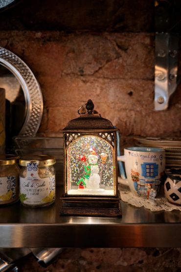 A festive snowman lantern surrounded by jars and mugs on a rustic shelf.