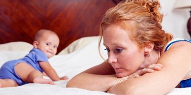 A woman lying on a bed with a baby crawling behind her.