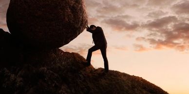 Person pushing a large boulder uphill at sunset.