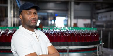 Man in a white shirt and cap stands confidently in a beverage production facility.