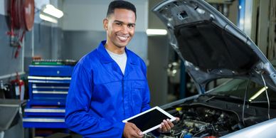 Smiling mechanic in blue uniform holding a tablet near a car with its hood open.