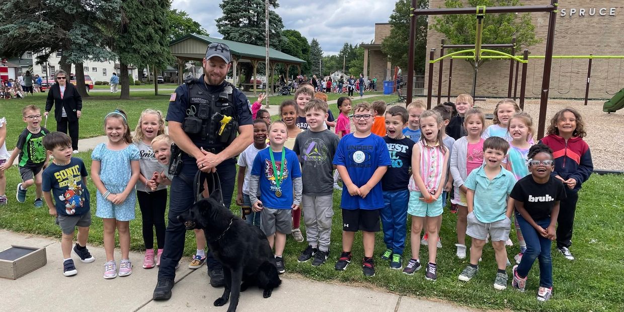 Police officer poses with smiling children and a black dog at a playground.