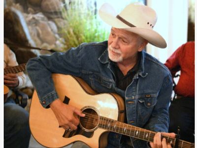 Older man playing acoustic guitar wearing a cowboy hat and denim jacket.