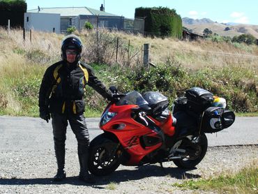 A motorcyclist in black and yellow gear stands next to a loaded red motorcycle on a rural road.