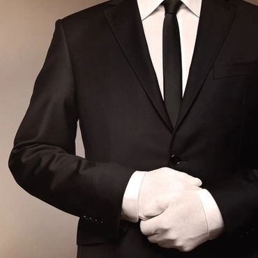 Man in black suit with white gloves, adjusting his cuff.