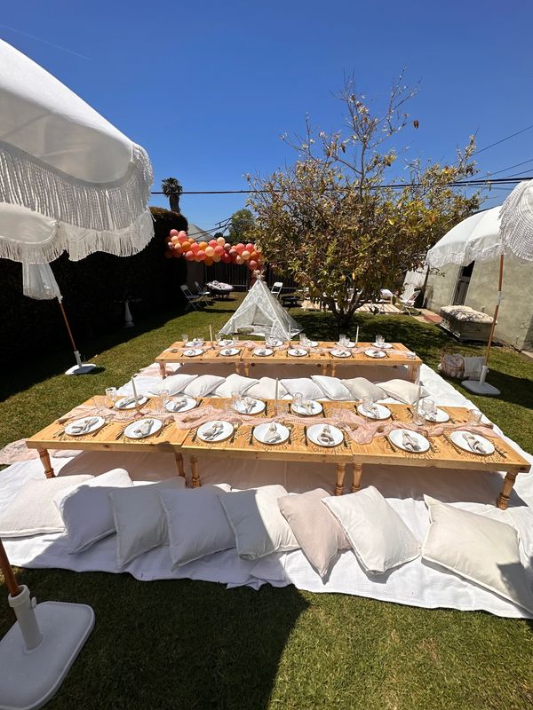 Outdoor picnic setup with low wooden tables, cushions, and decorative umbrellas on a sunny day.
