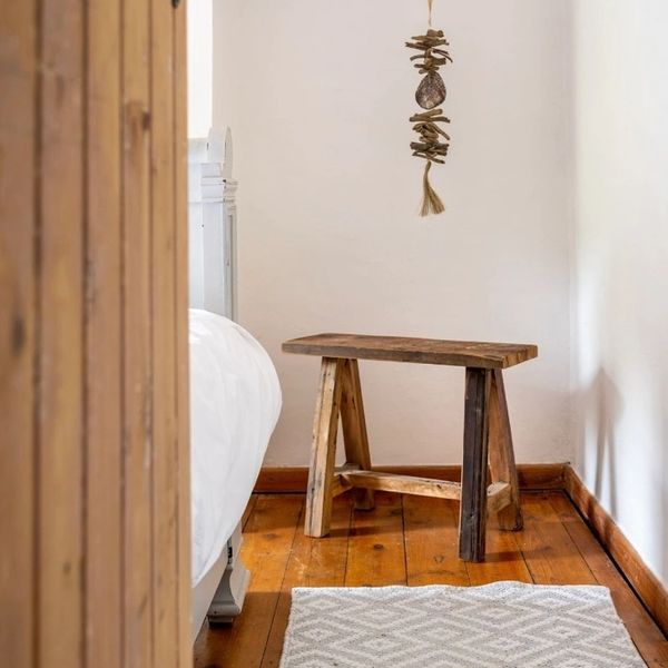 Cozy corner with rustic wooden stool and hanging decor beside a white bed.