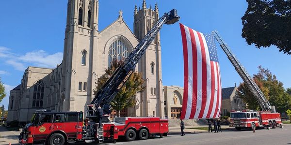 Two fire trucks hold a large American flag in front of a church.