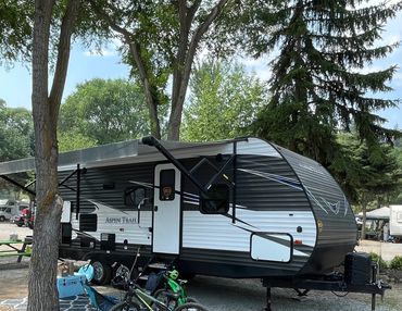 A parked Aspen Trail RV with bikes and camping gear nearby under trees.