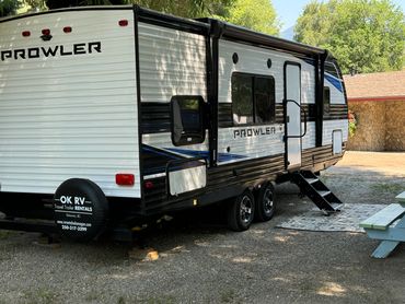 A Prowler travel trailer parked on gravel with stairs and picnic table nearby.