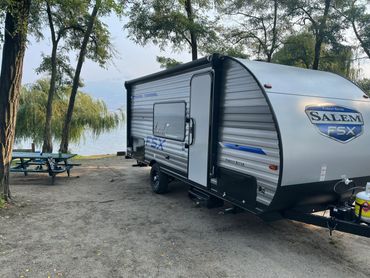 A Salem FSX travel trailer parked near a lakeside picnic area with trees.