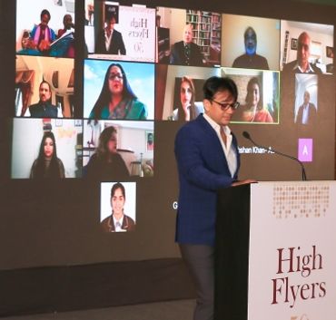 A man in a blue blazer speaks at a podium with a virtual meeting displayed behind him.